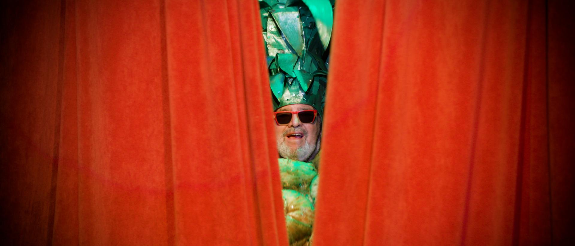 still from still pushing pineapples featuring a man wearing a pineapple costume and sunglasses peeking out between red curtains