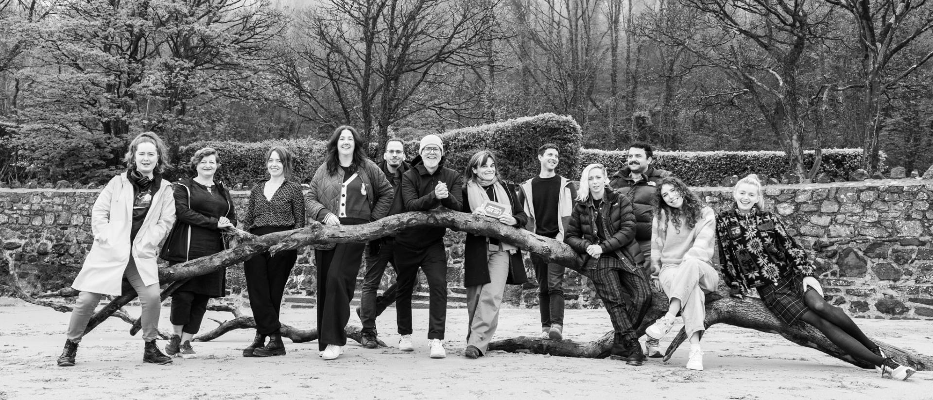 black and white photo of 12 people standing next to a fallen tree on a beach with a stone wall and forest behind them