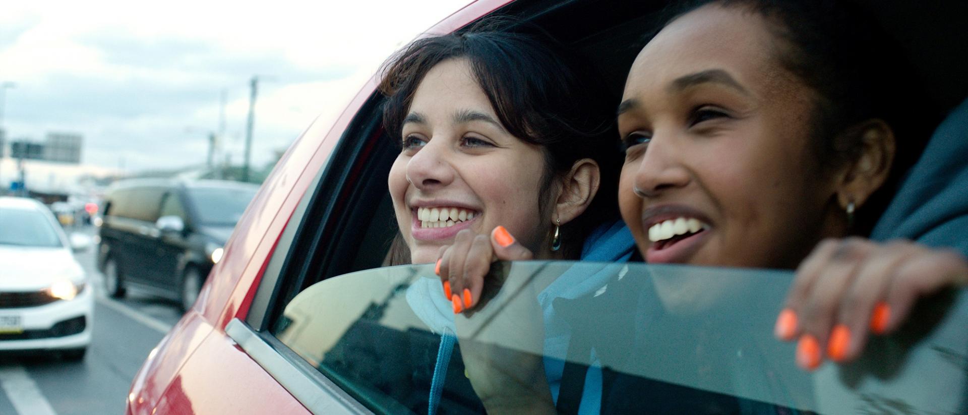 still from brides featuring two teenage girls in the backseat of a car peering out of the half-open window and smiling