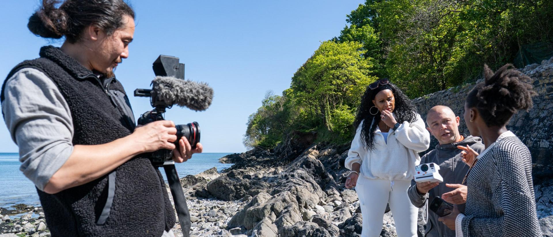 photo of four people on a pebble beach, one is filming the other three in conversation