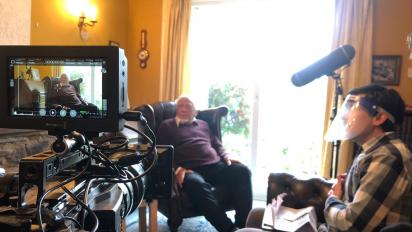 behind the scenes of a short film shoot, featuring a person being interviewed in their living room on camera. The interviewer is wearing a full face covering.