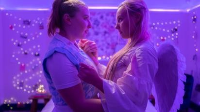 two women wearing party outfits beginning to embrace in a bedroom with fairy lights on the wall in the background