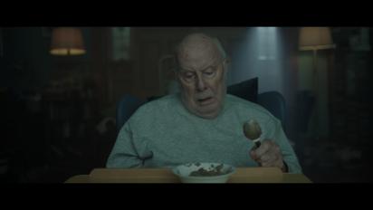 an elderly man sitting in a hospital chair looking down at a bowl of porridge
