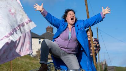 still from chuck chuck baby featuring a woman wearing a blue factory coat standing in front of a washing line and singing with her arms outstretched 