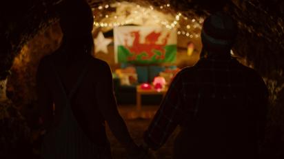 still from jelly featuring two people standing in a cave and holding hands while they look at a welsh flag decorated with fairy lights