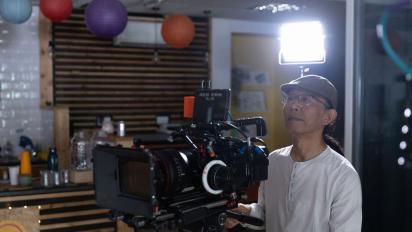 Photo of Keefa Chan standing in a community arts space, operating a camera a on a tripod with a light behind him. He is wearing a white top, a brown flat cap and glasses.