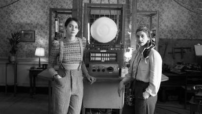 black and white still from lola featuring two people in 1940s costume standing next to their invention