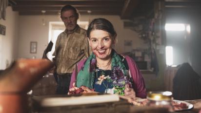 a woman sitting at a farmhouse kitchen table grinning creepily at the camera. A man stands behind her,