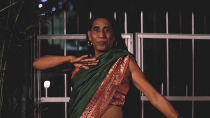 Still from India's First Best Trans Model Agency featuring a person wearing a green and red sari and posing for a photo in front of some metal gates.