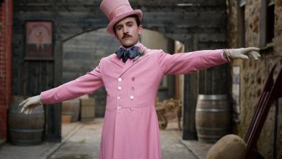 Still from Madfabulous featuring Callum Scott Howells wearing a pink suit and top hat and standing in a 19th century street. There are wooden barrels in the background.