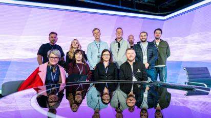 11 people sitting and standing around a reflective table in a TV news studio. 