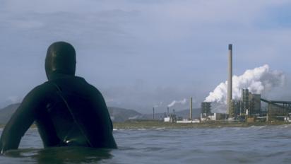 still from documentary smoking shores featuring a surfer in a wetsuit floating in the sea and looking at a steel factory in the background.