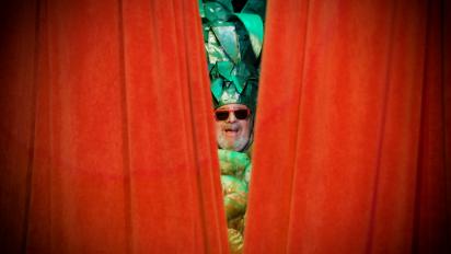 still from still pushing pineapples featuring a man wearing a pineapple costume and sunglasses peeking out between red curtains
