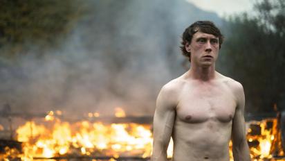still from mission featuring a topless man standing in front of a fire with a forest in the background
