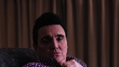 Still from short documentary film Juan, featuring a man wearing a purple shirt sitting at a white digital piano and looking into a wall mirror. .