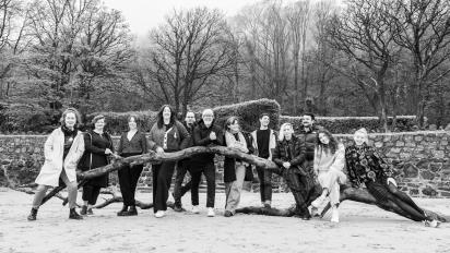black and white photo of 12 people standing next to a fallen tree on a beach with a stone wall and forest behind them