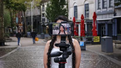 photo of a person standing in a city street in front of a tripod with a smartphone filming them